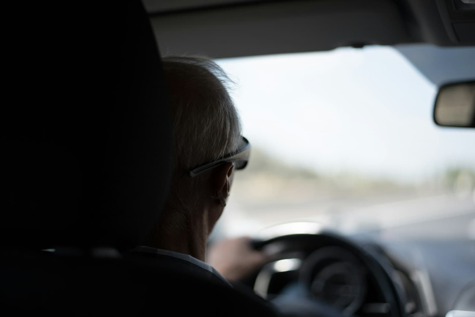 Elderly man in glasses driving in bright daylight, viewed from behind the driver's seat