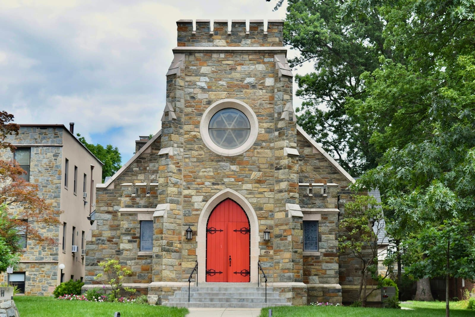 Stone church with red door, round window, and castellated tower surrounded by green trees