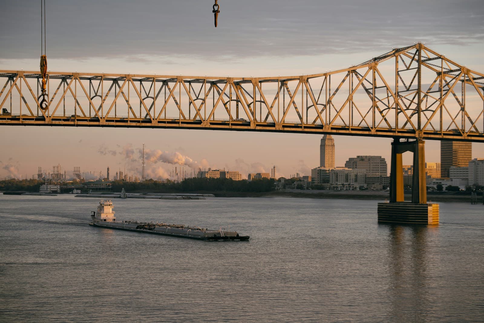 Steel truss bridge over river at sunset with barge passing underneath and city skyline in background
