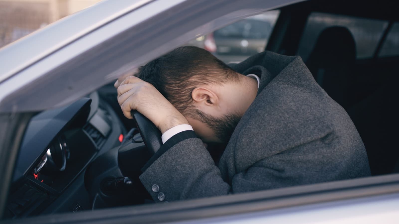 Man in a dark suit resting his forehead on the steering wheel in distress
