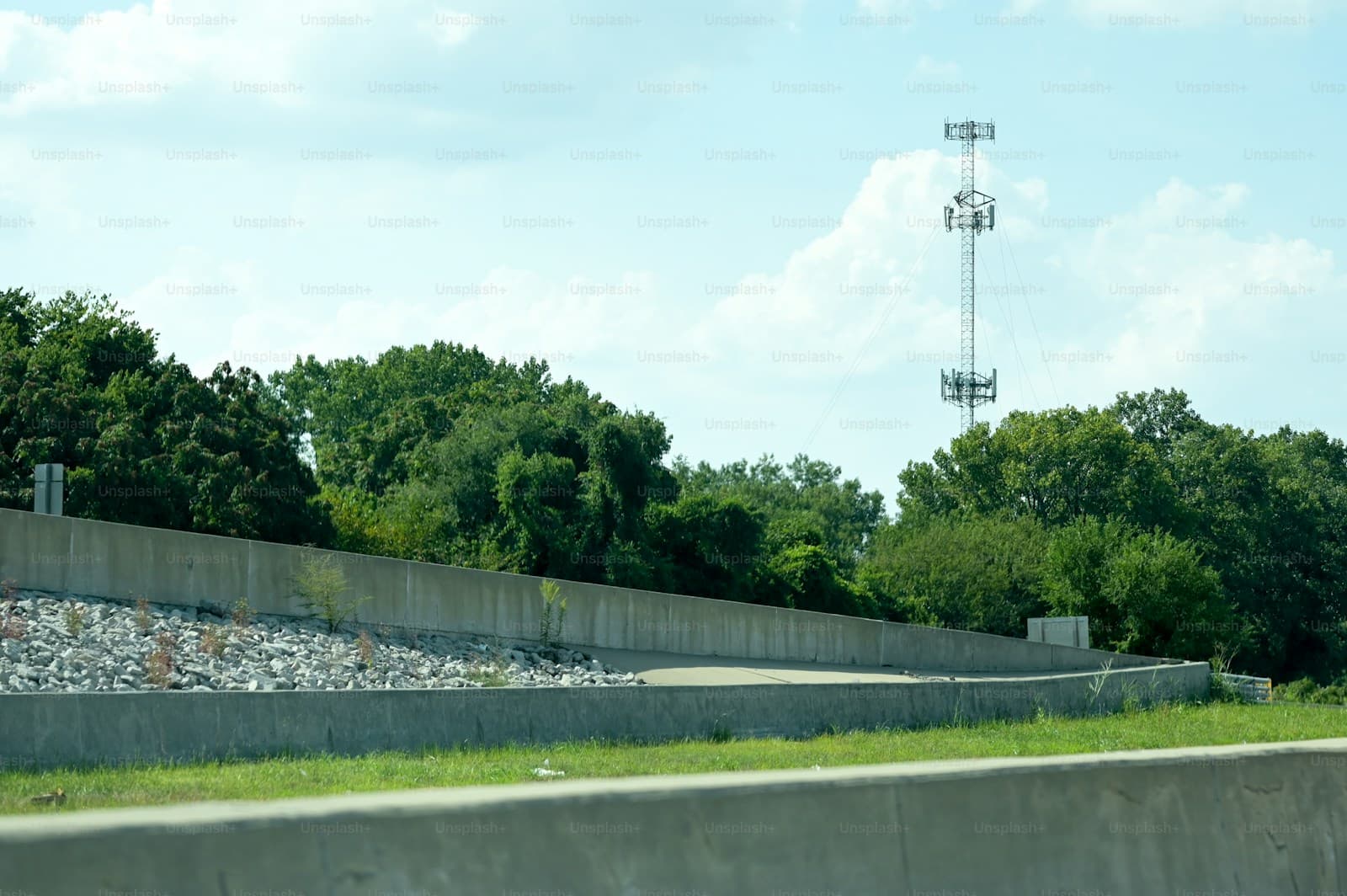Concrete retaining wall along road with cell tower visible behind trees against cloudy sky