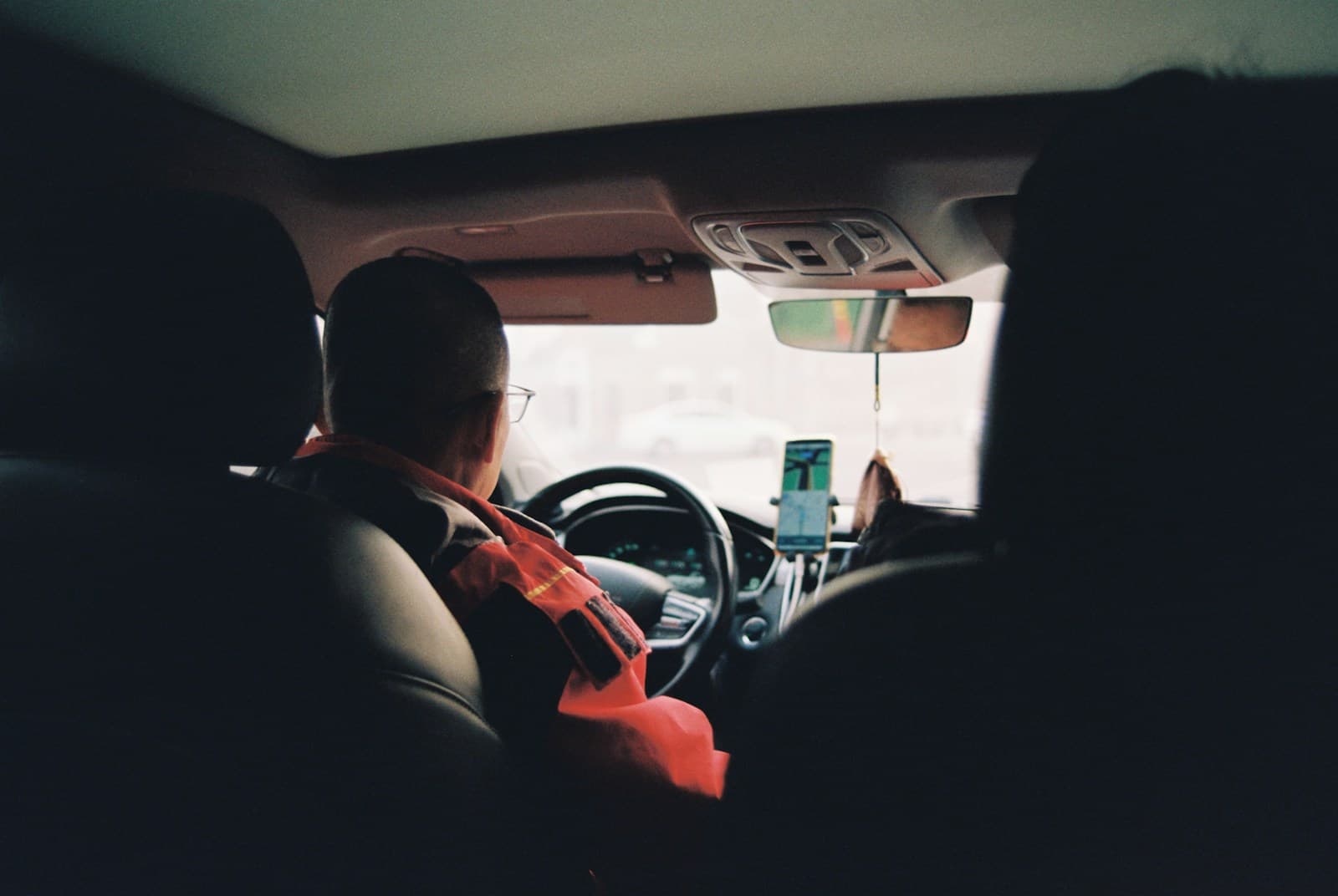 Driver in an orange jacket at the wheel with a phone mount on the dash, seen from the back seat