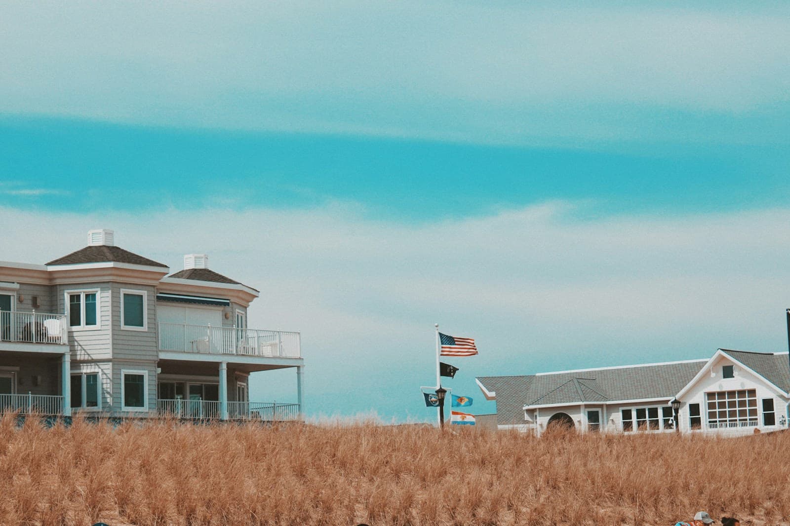 Two coastal beach houses with American flag flying between them, surrounded by golden marsh grass under blue sky