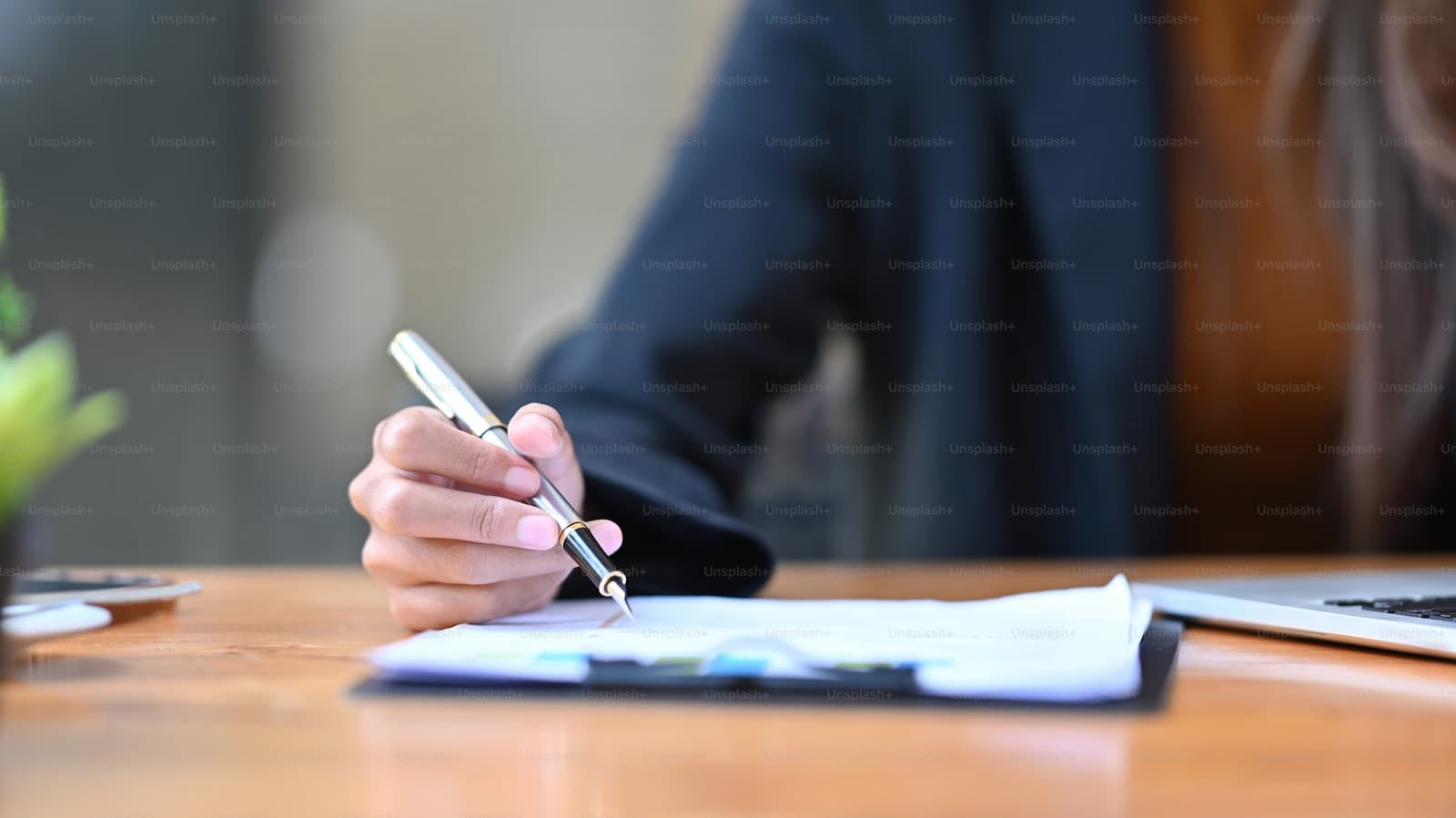 Person in business attire writing with pen on documents at wooden desk in office setting