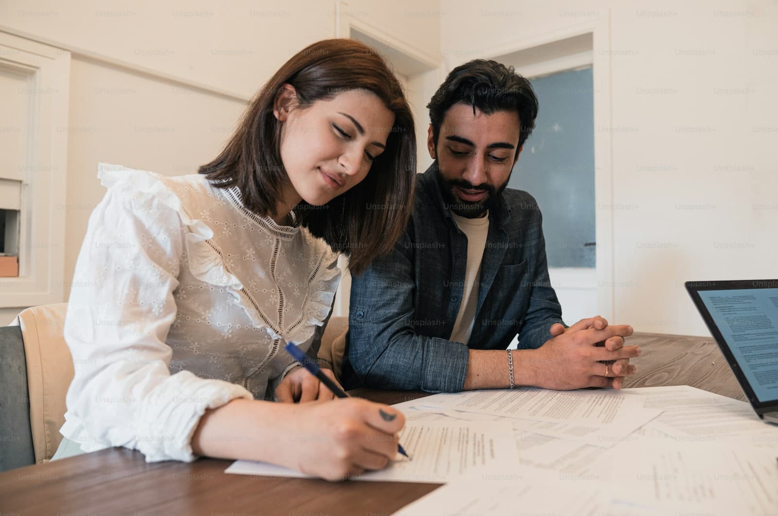 Young woman signing documents at a table as a bearded man watches and smiles