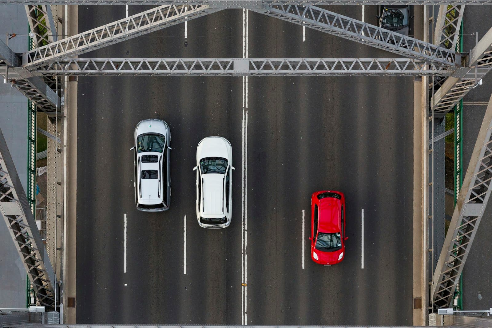 Aerial view of three cars on a steel truss bridge - two white cars and one red car driving in separate lanes