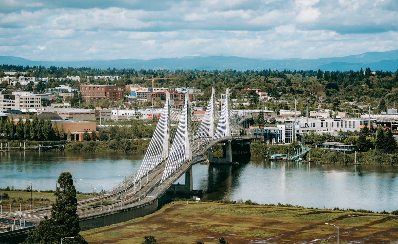 Aerial view of white cable-stayed bridge crossing river with city buildings and mountains in background