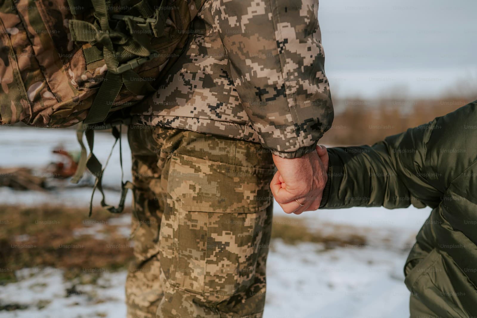 Two people in camouflage uniforms holding hands in the snow, one with a military pack