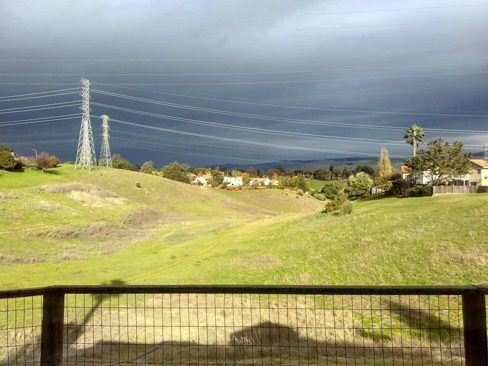 Rolling green hills under stormy skies with power lines, houses, and palm trees in the distance