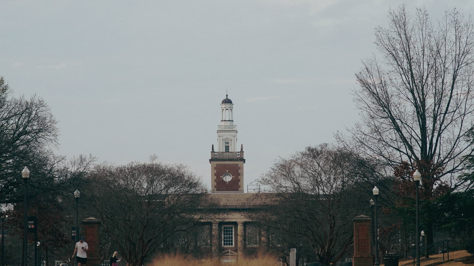 Historic brick building with white clock tower surrounded by bare trees under overcast sky