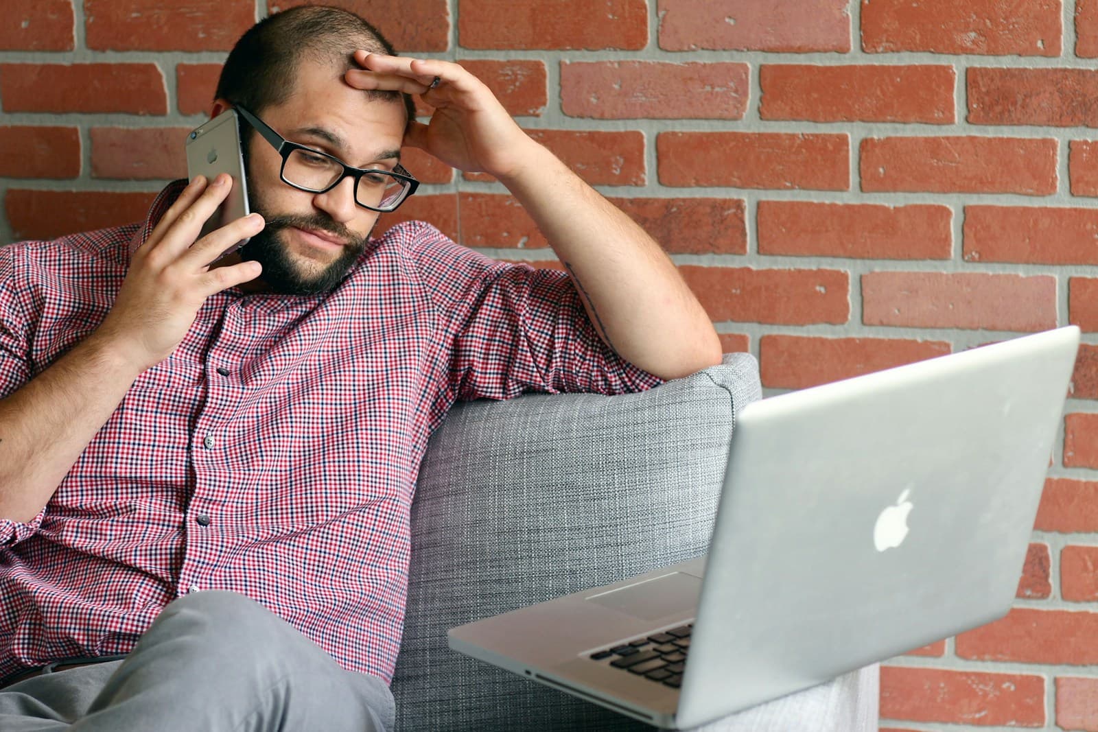 Man in a checkered shirt on a couch holding his head while on the phone with a laptop open