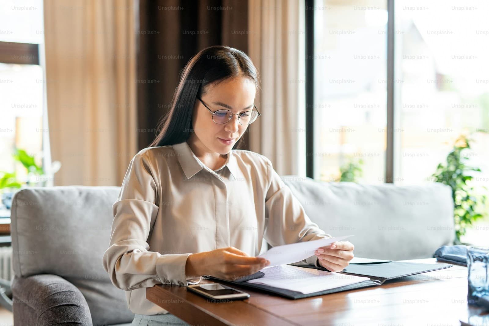 Professional woman in glasses and beige shirt reviewing documents at wooden table in bright home office setting
