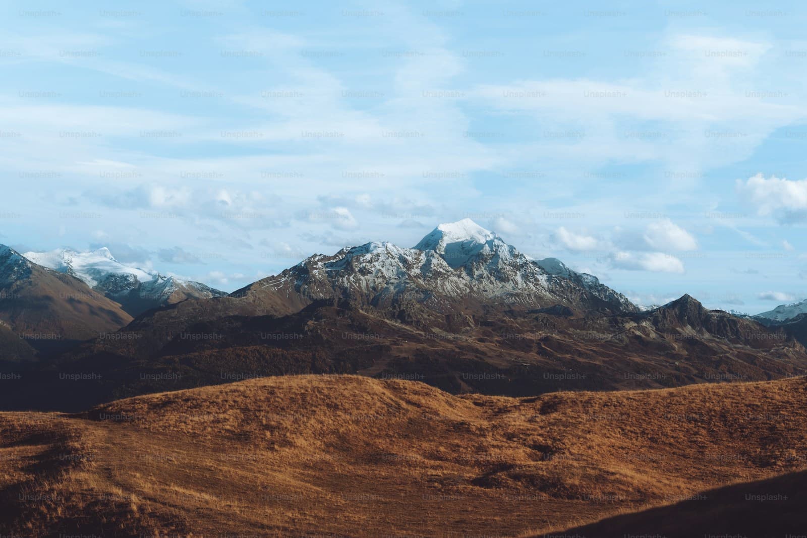 Snow-capped mountains rising behind golden-brown rolling hills under a cloudy blue sky