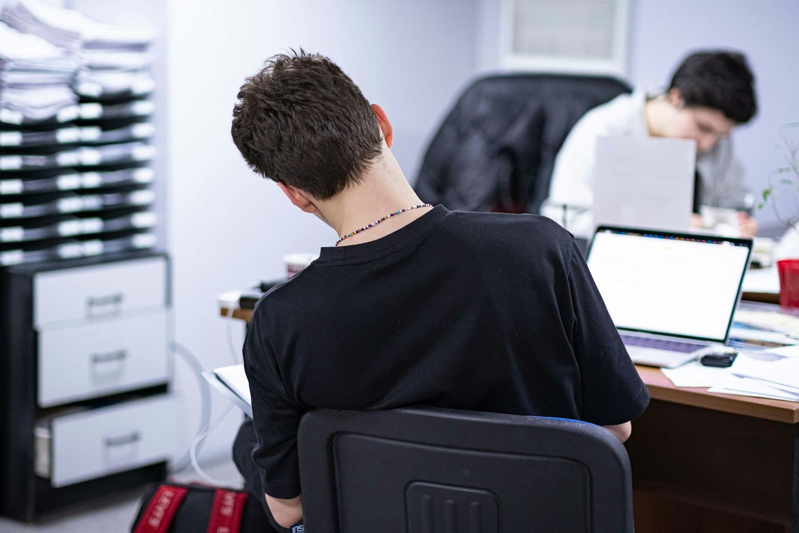 Man in black shirt working on laptop at office desk with female colleague in background