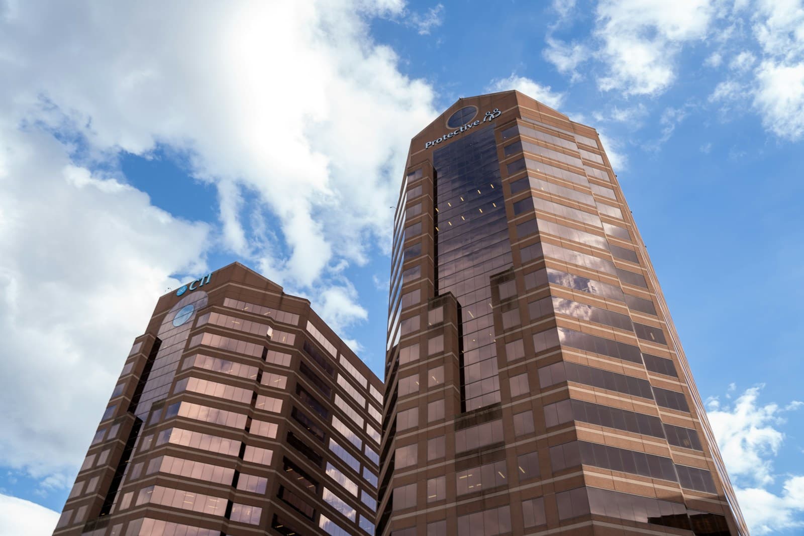 Two tall modern office buildings with reflective glass facades shot from below against a cloudy blue sky