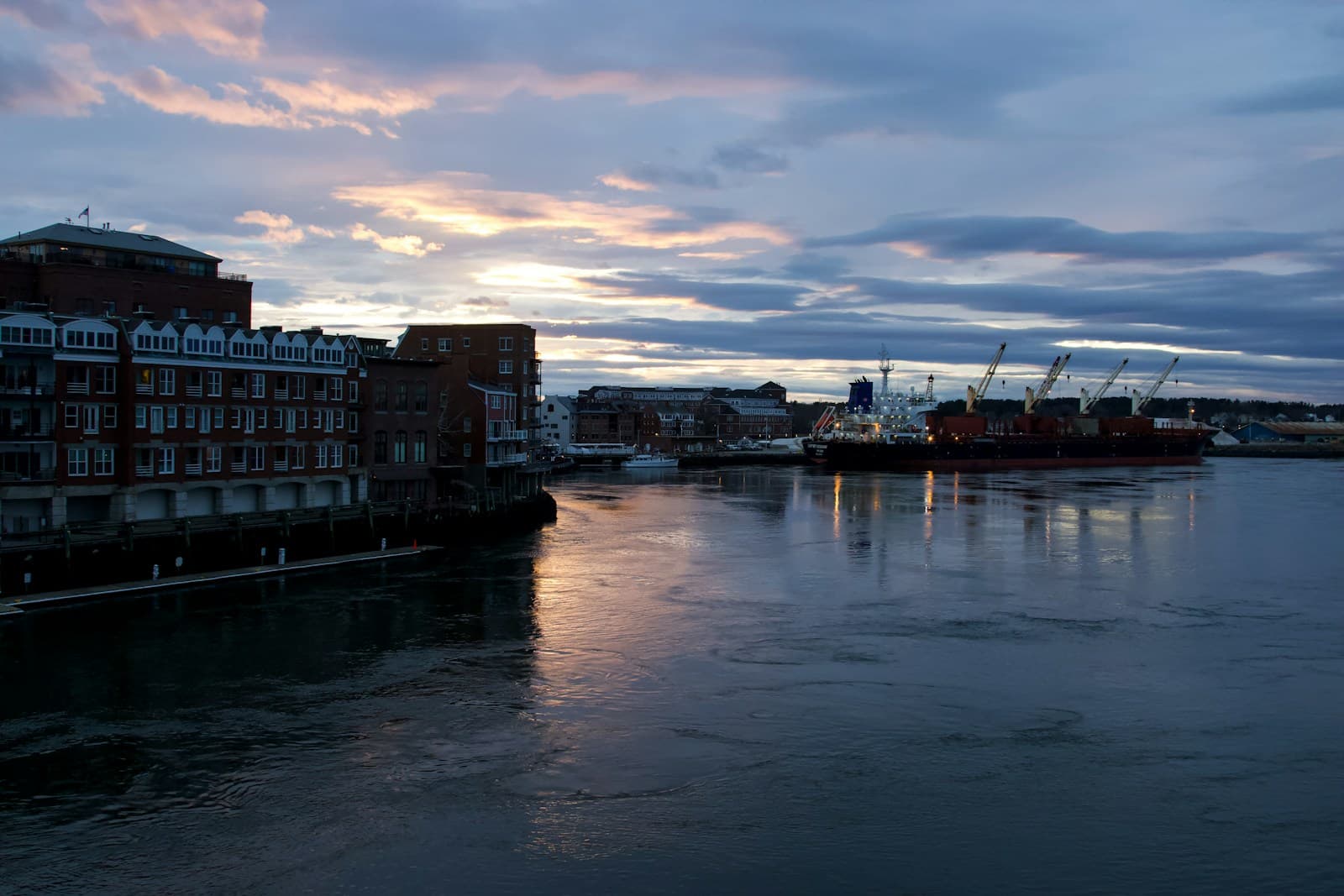Waterfront city view at sunset with brick buildings reflected in calm water under dramatic cloudy sky