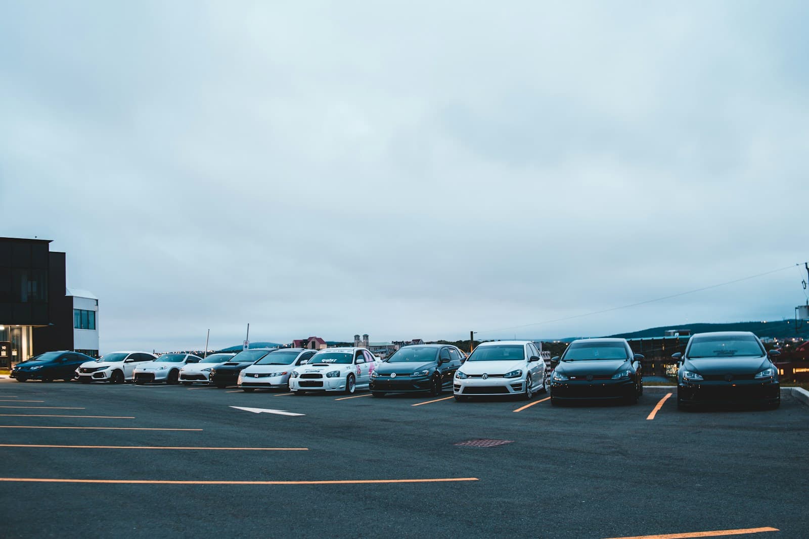 Cars parked in rows in a large parking lot during twilight with overcast sky and buildings in background