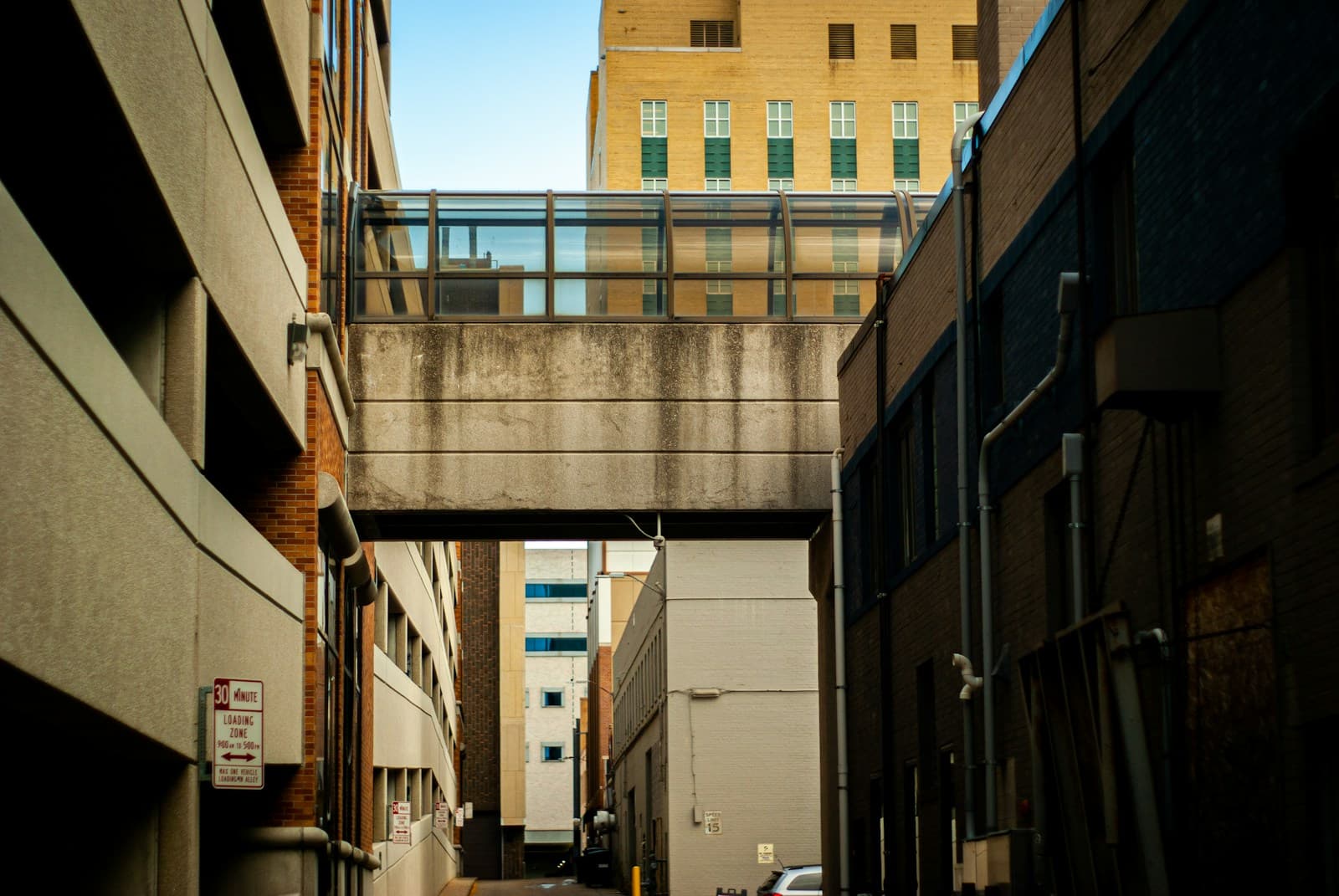 Urban alley with glass pedestrian bridge connecting buildings overhead, surrounded by brick and concrete architecture