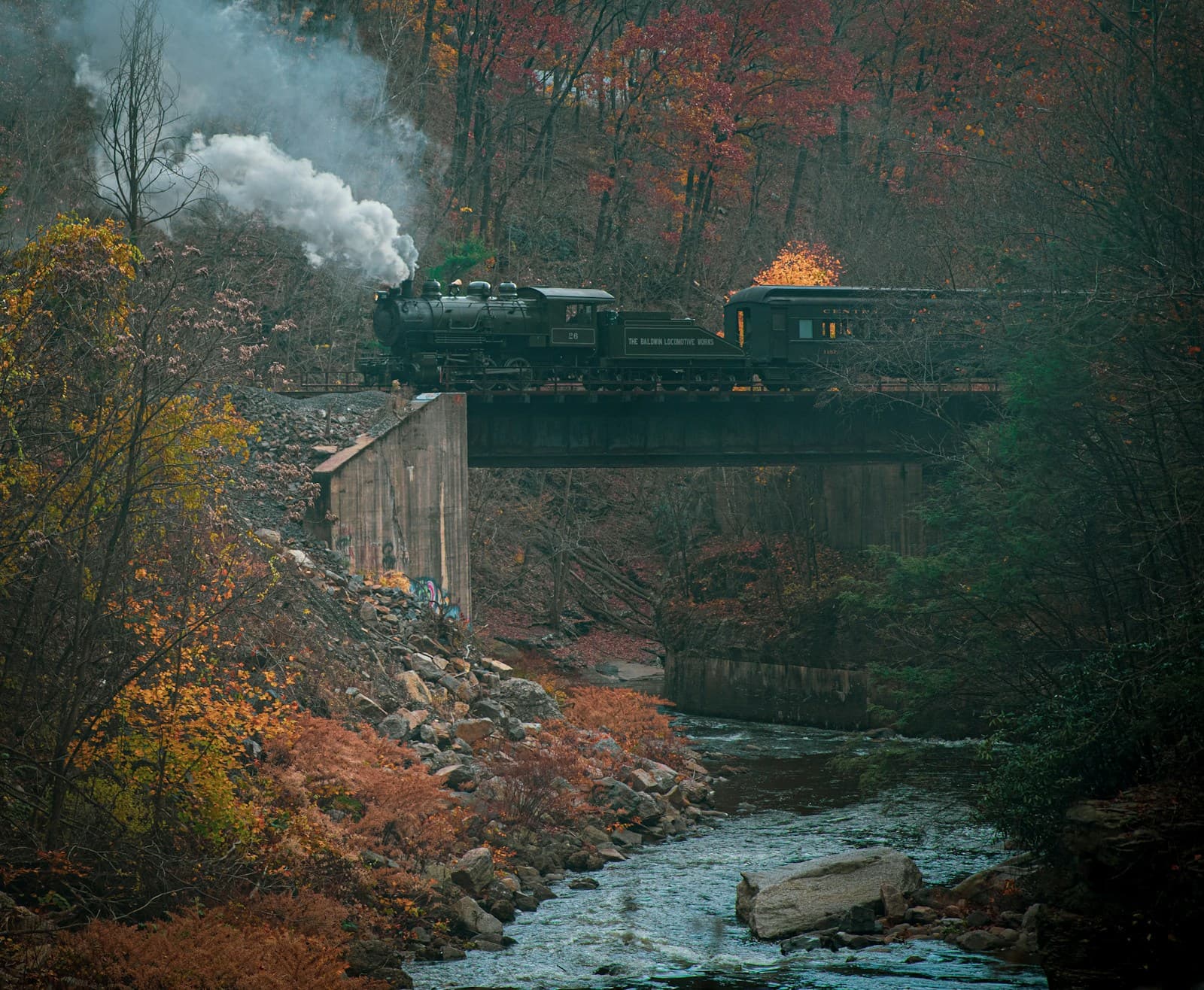 Steam train crossing stone bridge over creek in autumn with colorful fall foliage and billowing white smoke
