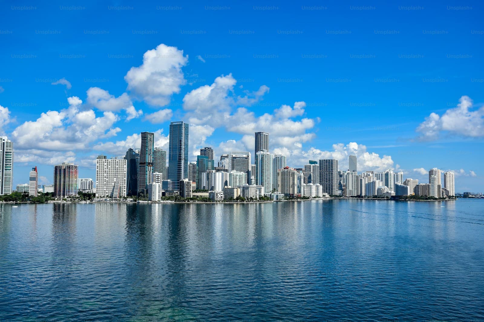 Modern city skyline with tall buildings reflected in calm blue water under a bright sky with white clouds