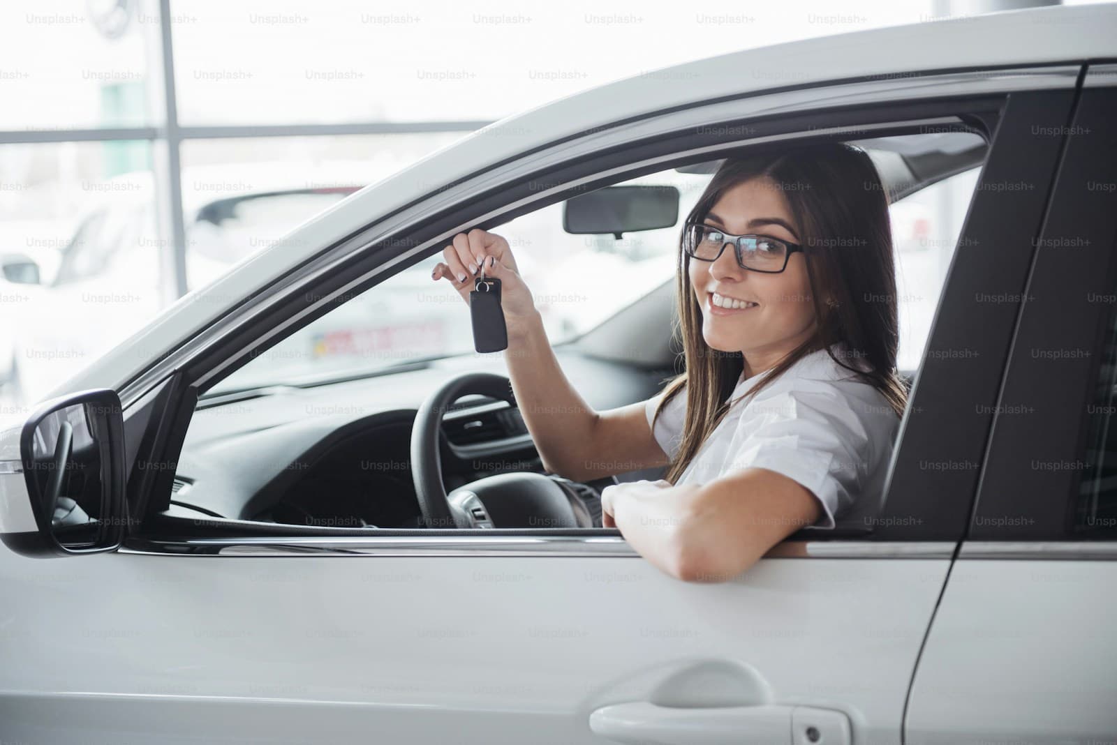 Young woman in glasses holding up car keys in the driver's seat of a white car