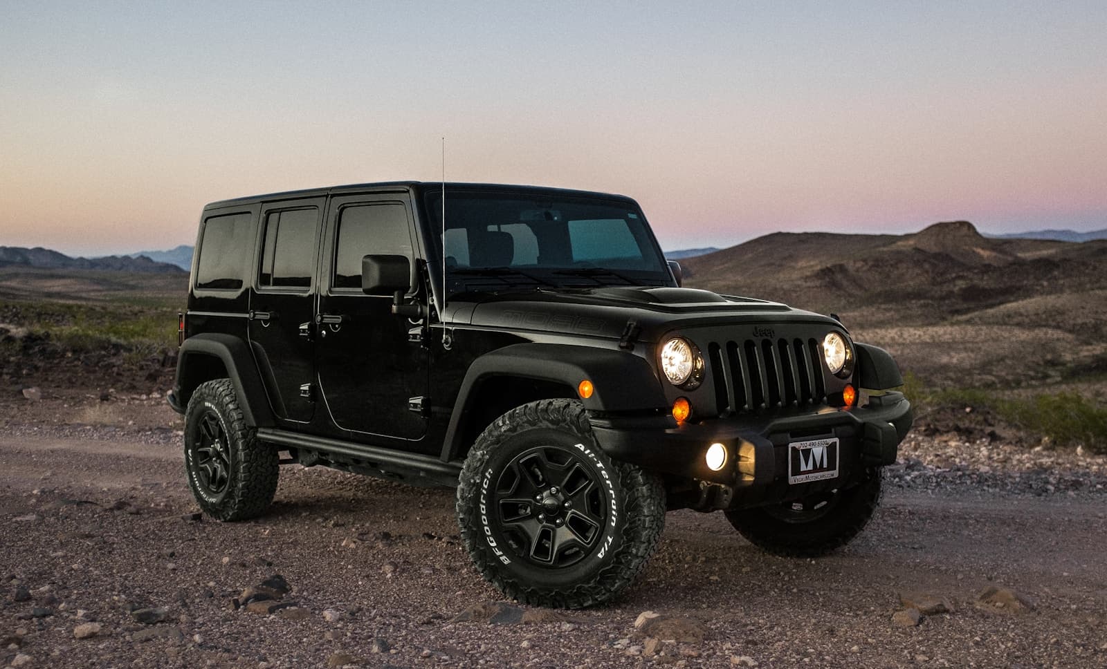 Black Jeep Wrangler parked on rocky desert terrain at sunset with headlights on
