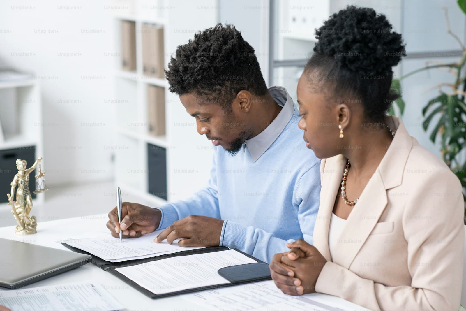 Black man signing documents while Black woman in business attire watches in modern office setting