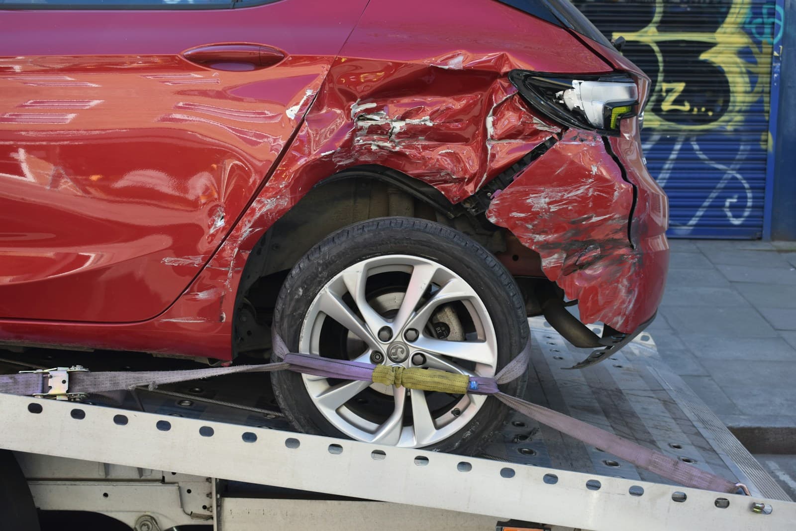 Red car with severe rear quarter-panel and wheel-well damage on a loading ramp