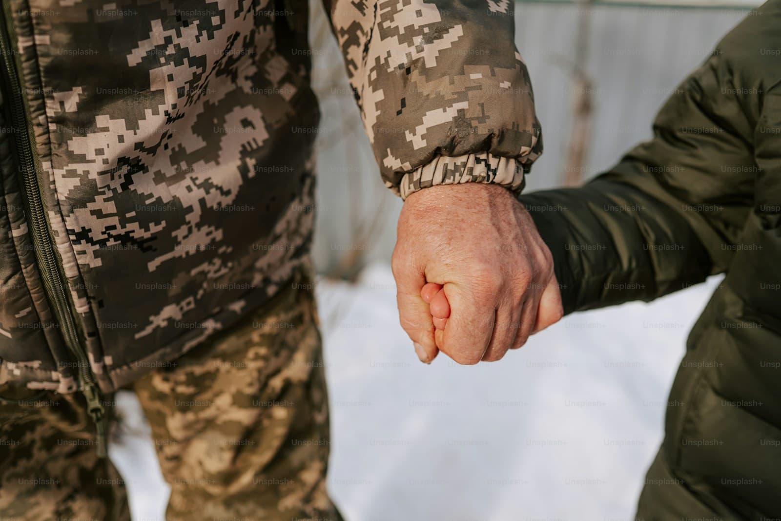 Two people in camouflage uniforms holding hands, one in an olive green jacket