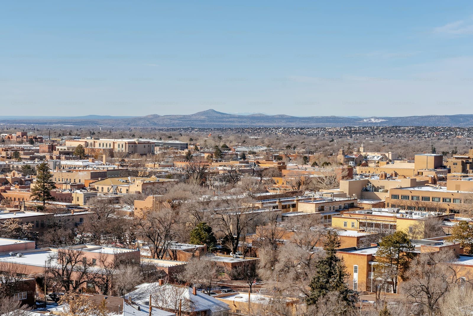 Aerial view of city skyline with buildings, bare trees, snow patches, and mountains in background under blue sky