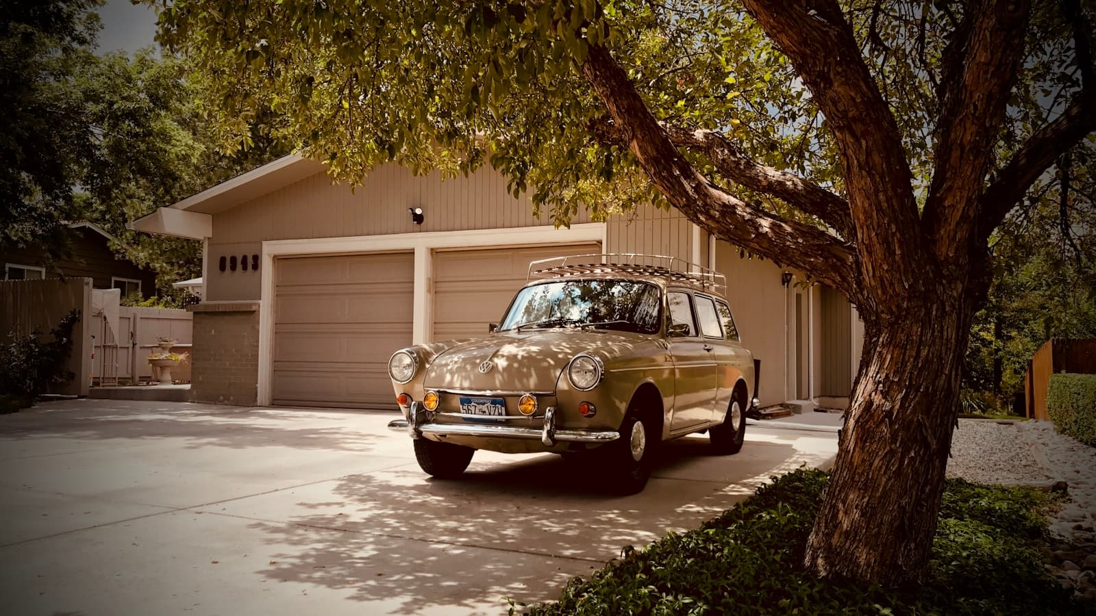 Vintage tan car parked in driveway under large tree with suburban house and garage in background