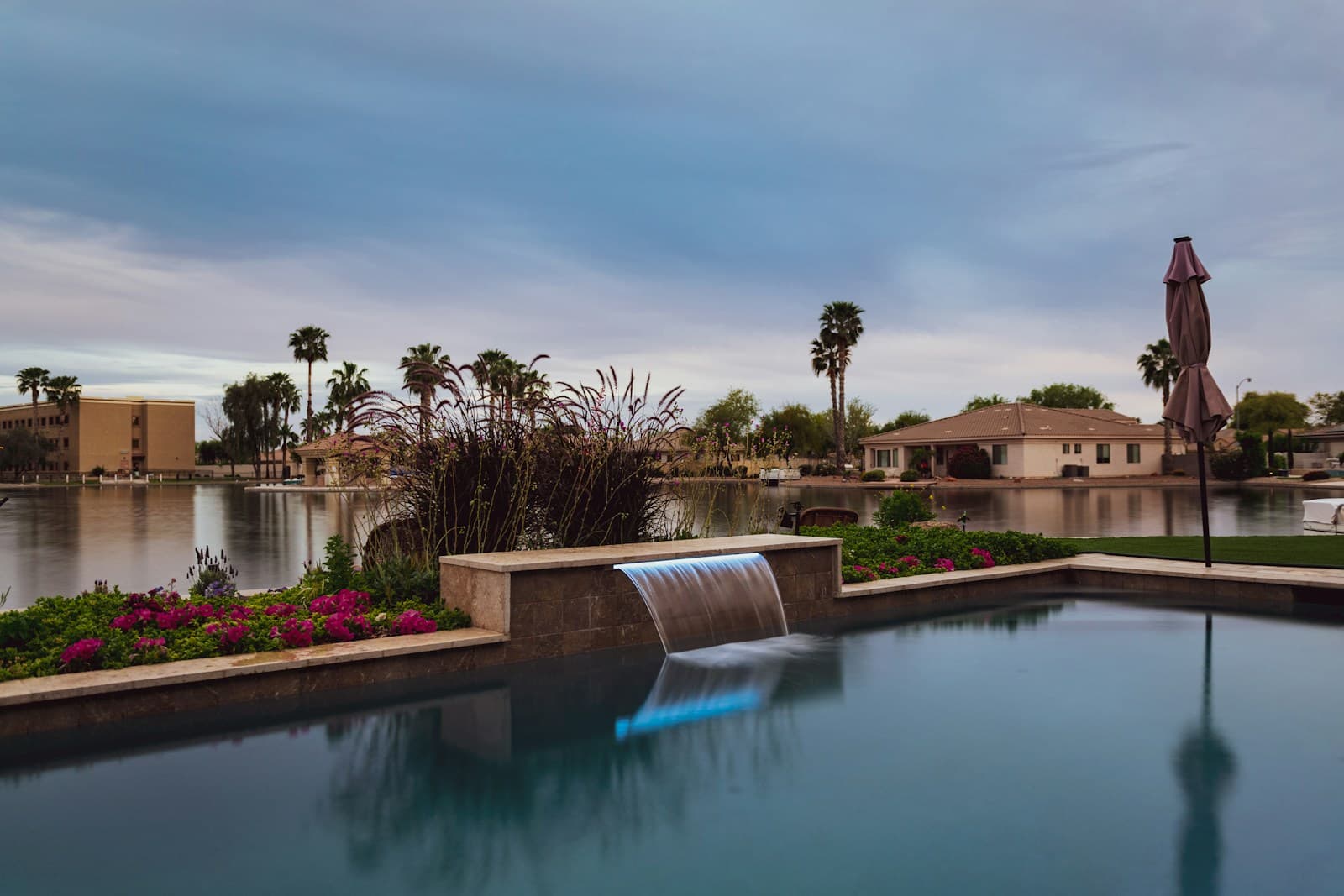 Modern infinity pool with waterfall feature overlooking lakefront homes and palm trees at twilight