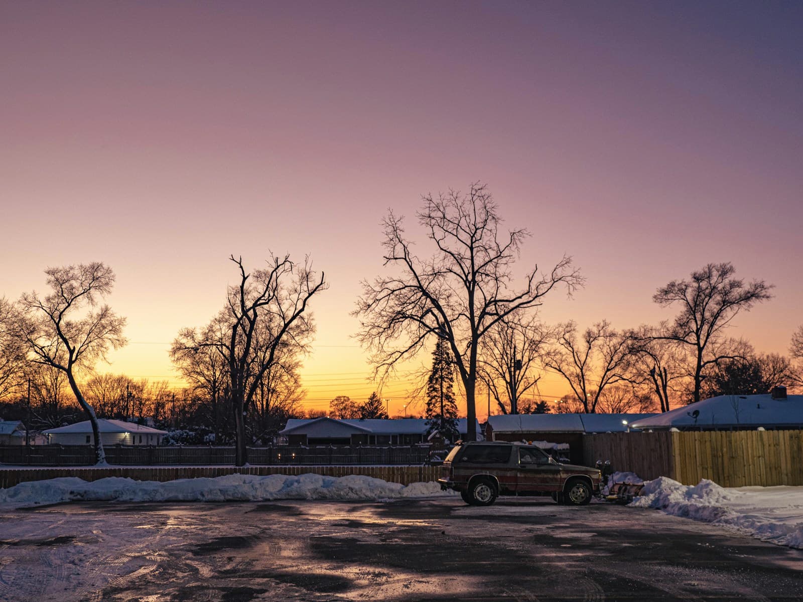 Pickup truck in snowy parking lot at sunset with bare trees and purple-orange sky in winter suburban setting