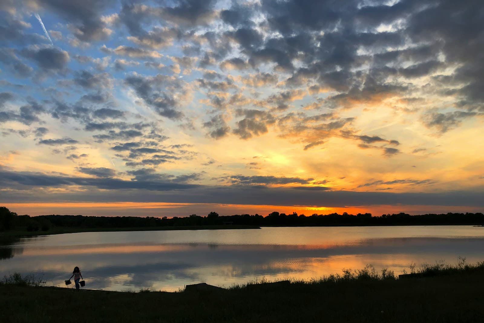 Sunset over calm lake with dramatic cloudy sky reflections and silhouette of person sitting by water