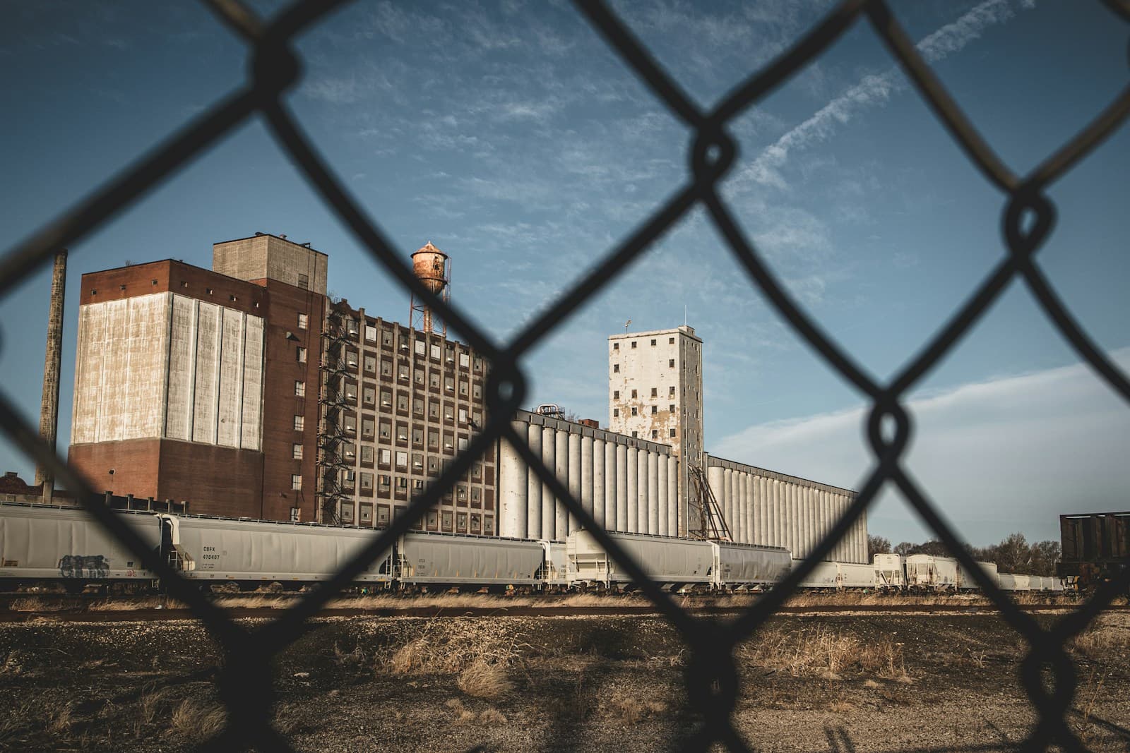 Industrial buildings and grain silos viewed through chain-link fence under cloudy sky