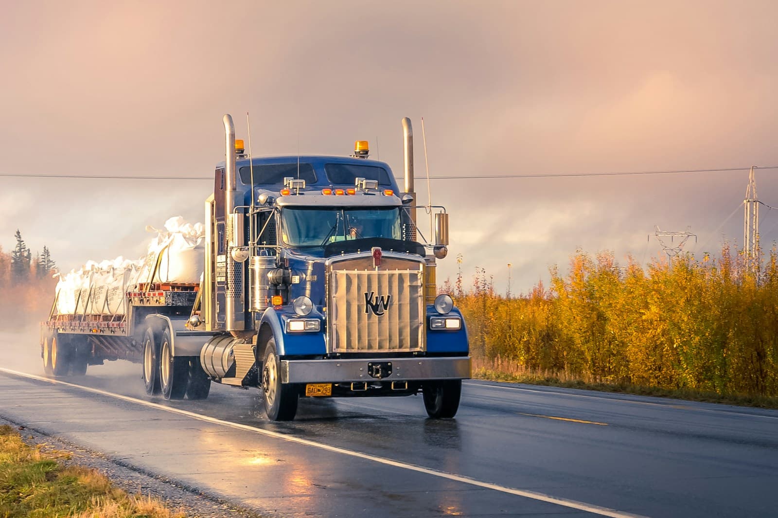 Blue semi-truck with chrome stacks on a highway at sunset amid autumn foliage