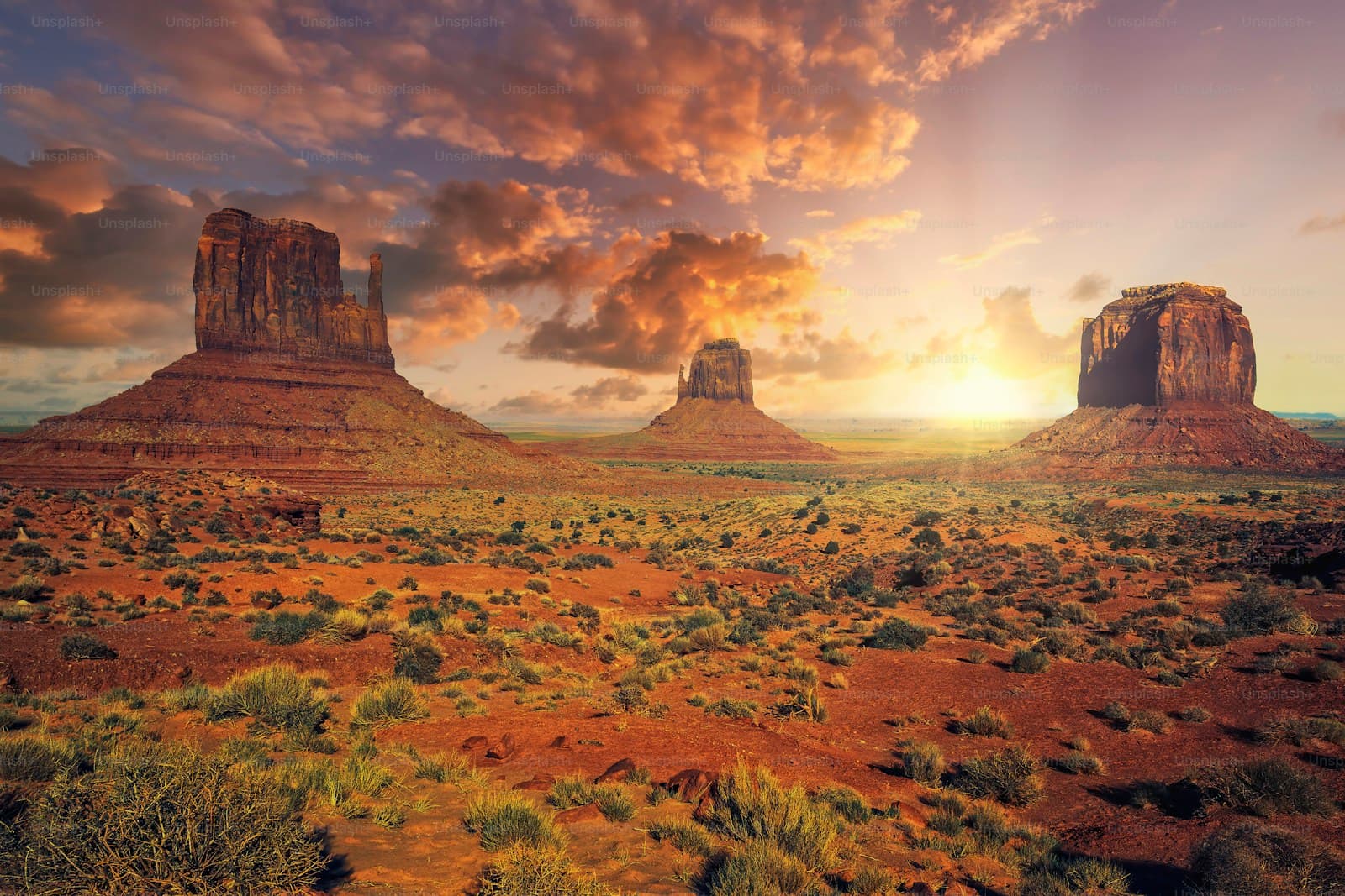 Desert landscape with red rock formations and dramatic sunset sky over sparse vegetation and red soil