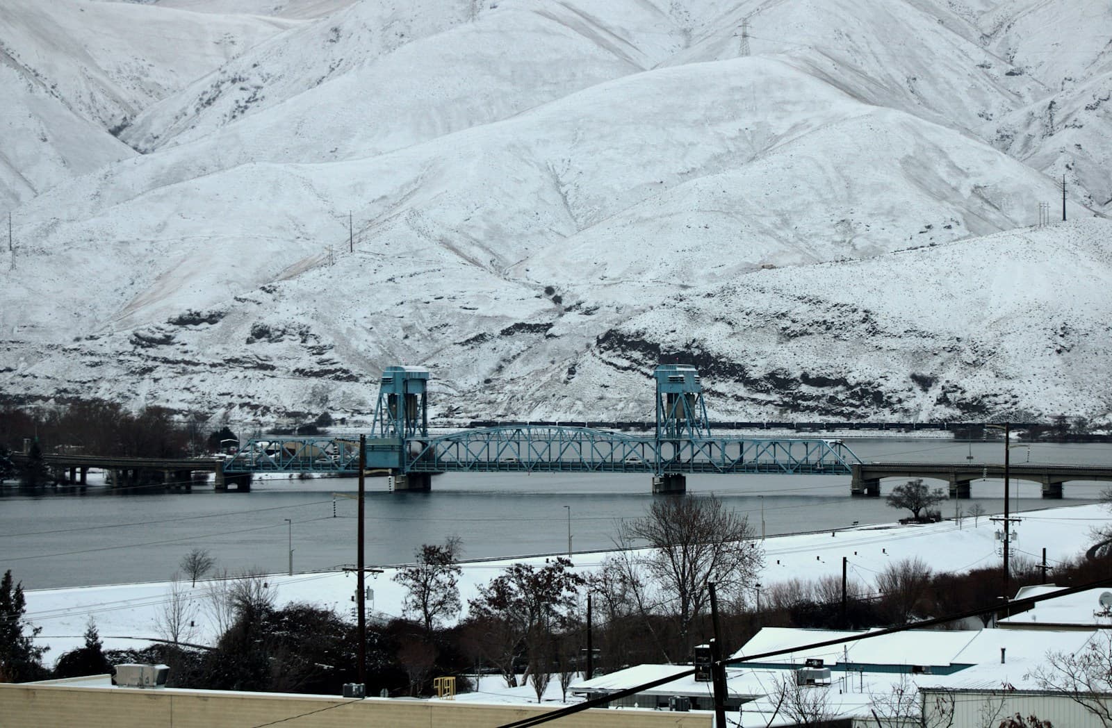 Green steel bridge crossing water with snow-covered mountains and hills in background during winter