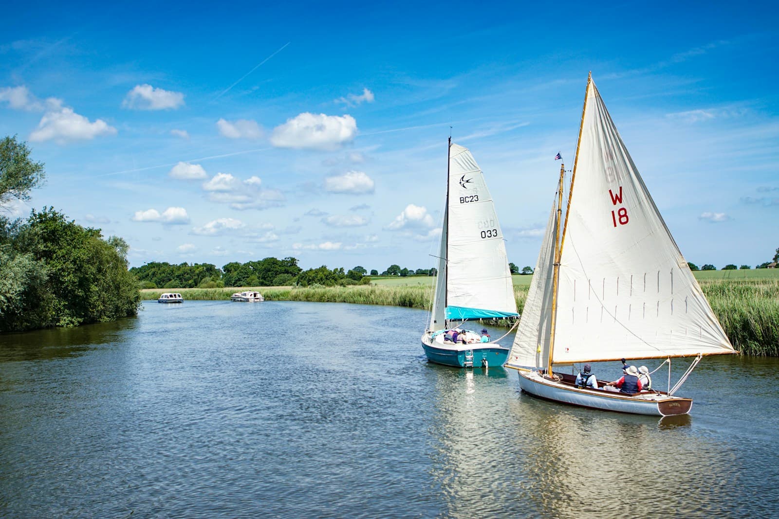 Two white-sailed boats on a calm river with green countryside and blue sky in background