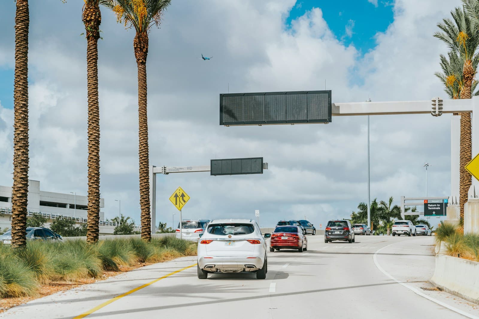Cars driving on a multi-lane road with palm trees and traffic signals overhead under partly cloudy skies