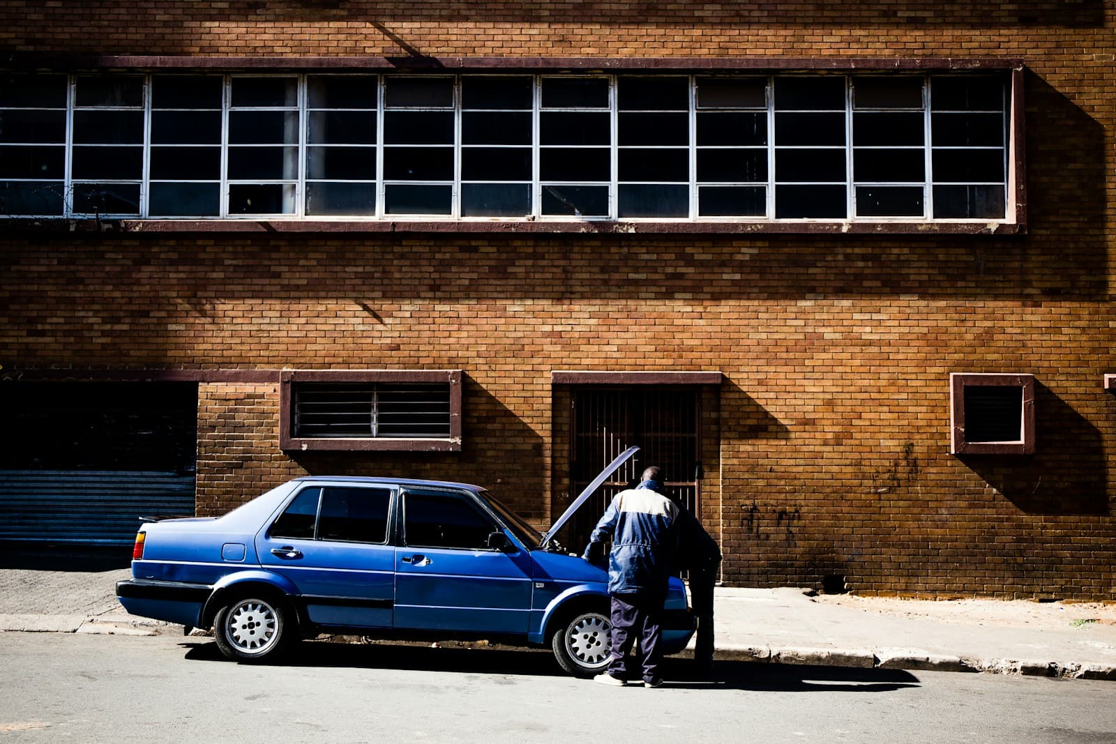 Person standing beside a blue sedan parked in front of a brick industrial building