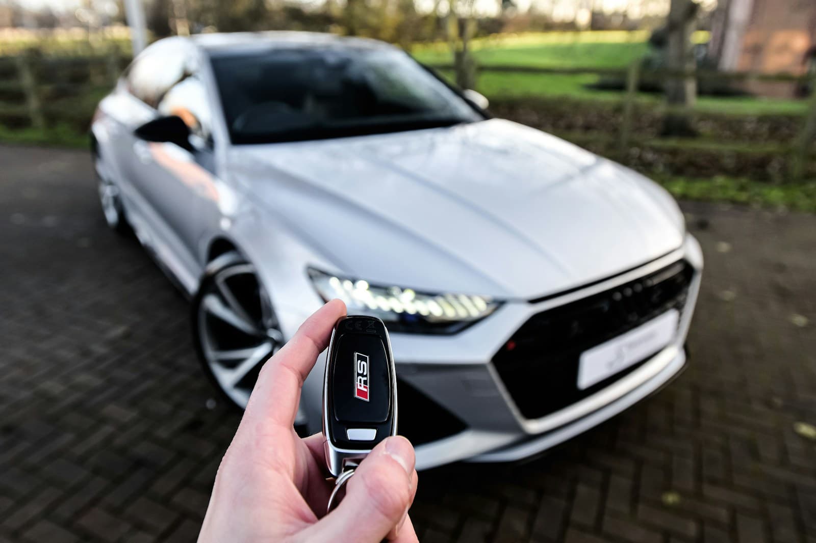 Hand holding a key fob in front of a white luxury sedan on a residential street