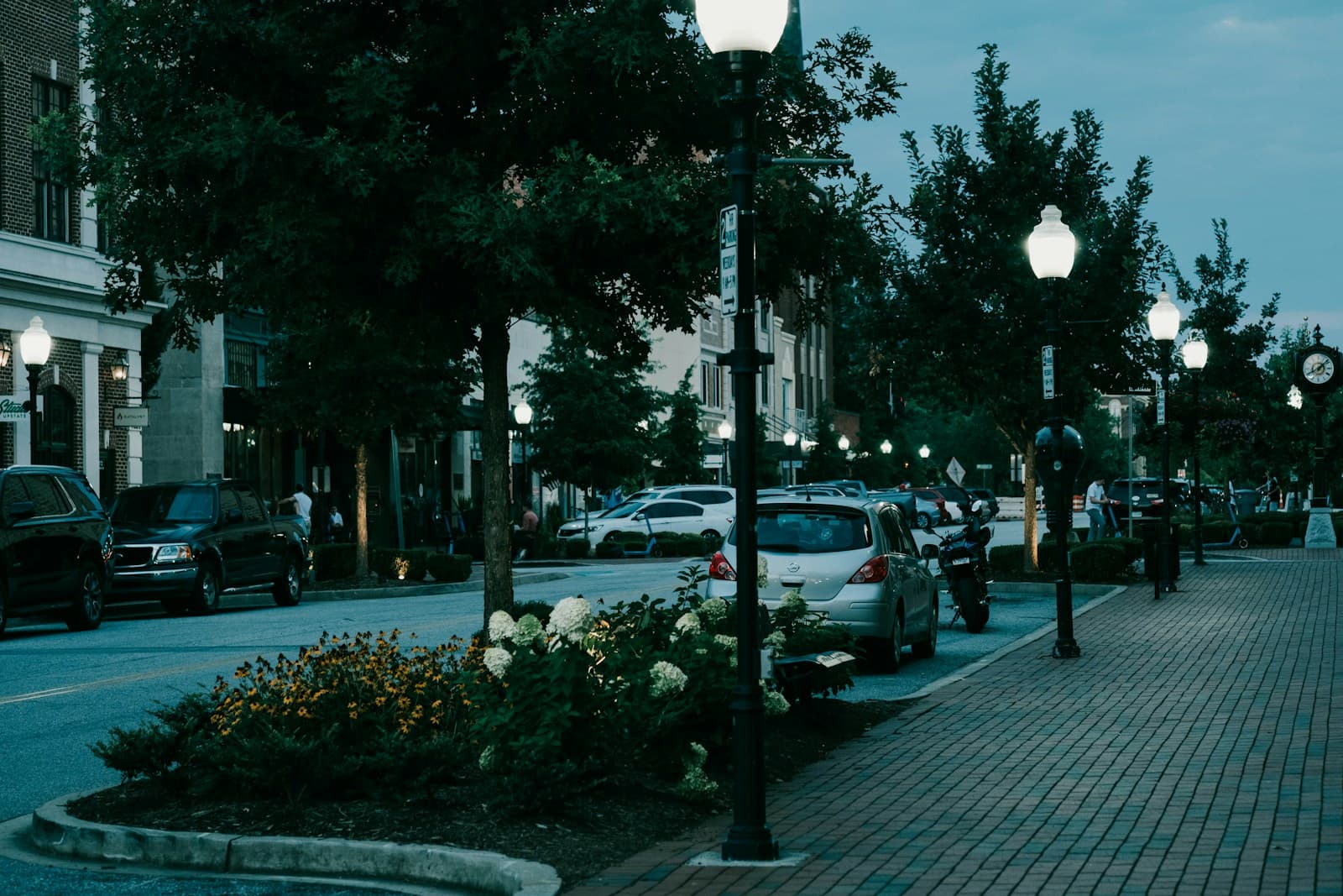 Evening street scene with glowing street lamps, parked cars, and landscaped flower beds in downtown area