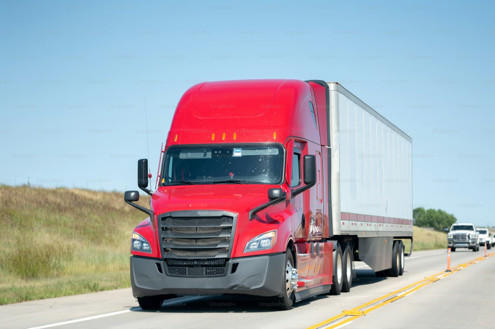 Red semi-truck with white trailer driving on rural highway under blue sky